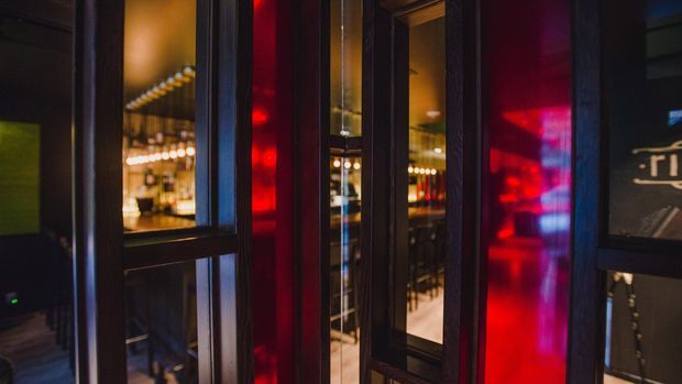 A moody close-up view through dark-framed glazed partitions or doors into a warmly lit bar or hospitality interior. Deep red illuminated panels and reflections contrast with black frames, while bottles, shelving, pendant lights, and bar seating are visible beyond the glazing.