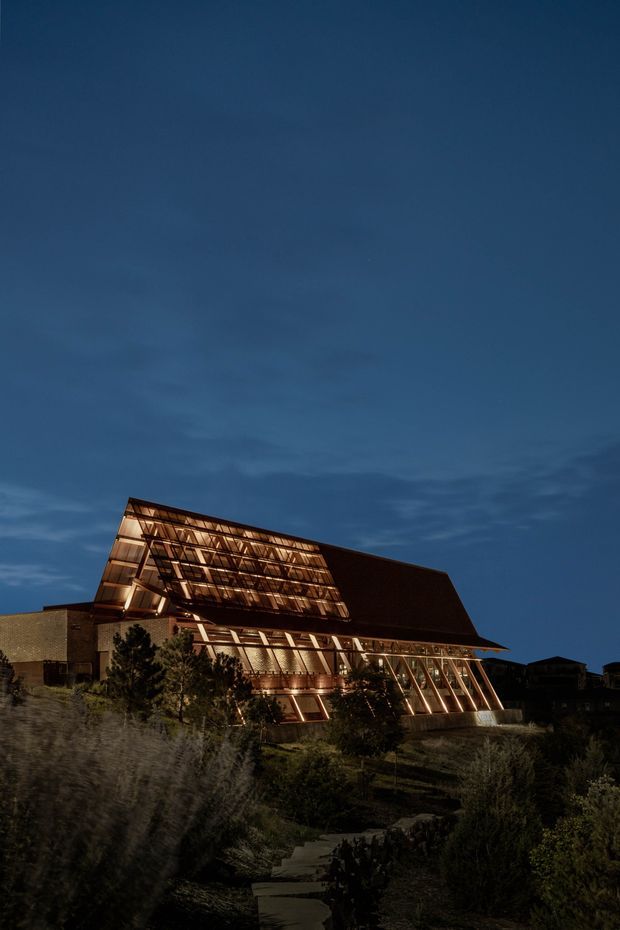 A dramatic twilight exterior photograph of a large contemporary building set on a landscaped, sloping site. The structure features extensive warm-lit glazing, a strongly angular and cantilevered form, a steep dark roof plane, and prominent exposed structural framing. A planted pedestrian path leads toward the building through dense vegetation.