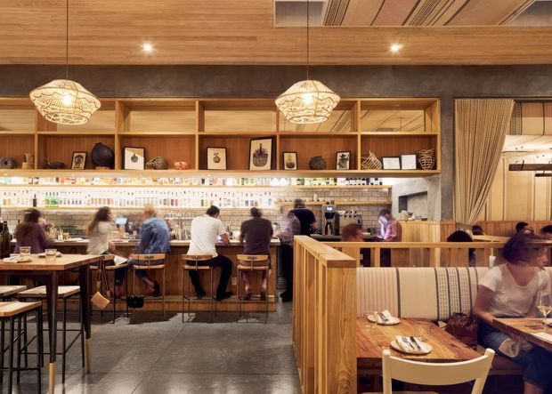 Warmly lit restaurant interior with a long bar and open service counter, timber shelving and wall panelling, decorative pendant lights, tiled backsplash, and booth seating separated by timber screens. Diners and bar patrons occupy the space, which has a refined contemporary hospitality design with natural timber and dark stone-like finishes.