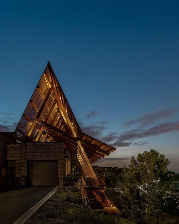 Twilight exterior view of a dramatic contemporary house on a sloped site. The residence features a sharply angled triangular roof and gabled form, extensive warm timber cladding and exposed structural elements, a dark brick garage volume, and illuminated timber-lined openings and deck areas. Landscaping and distant suburban terrain are visible beneath a deep blue evening sky.