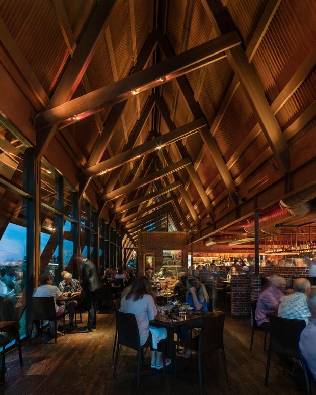 Interior photograph of a busy restaurant or hospitality venue with a dramatic vaulted timber roof and exposed heavy timber trusses. Warm amber lighting highlights the timber ceiling, dark wood flooring, exposed services, brick bar elements, and numerous dining tables. Floor-to-ceiling glazing lines one side of the space, with a bar and open dining area extending through the interior.