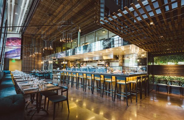 A sophisticated restaurant or hospitality interior with a long illuminated bar, high stools, dining tables, and booth seating. The space features exposed industrial-style services and ductwork, extensive glass, polished concrete flooring, dark timber slatted ceiling and overhead screens, suspended pendant lights, and a richly layered contemporary fit-out.