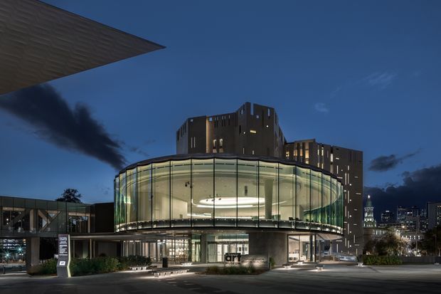 Night-time exterior view of a contemporary multi-storey civic or commercial building. The main feature is a large rounded glass-enclosed upper-level volume with a softly illuminated interior, supported by a concrete podium and glazed entrance below. Dark rectilinear tower forms rise behind it, while a broad projecting canopy frames the upper left. Urban buildings are visible in the background.