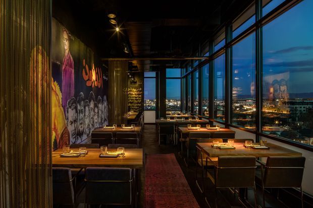 A dark, atmospheric high-rise restaurant or dining venue with rows of timber dining tables, upholstered chairs, warm pendant and recessed lighting, expansive floor-to-ceiling windows, and a nighttime city view. A dramatic graffiti-style mural and textured metallic curtain line one wall, while a long central runner adds warmth and depth.