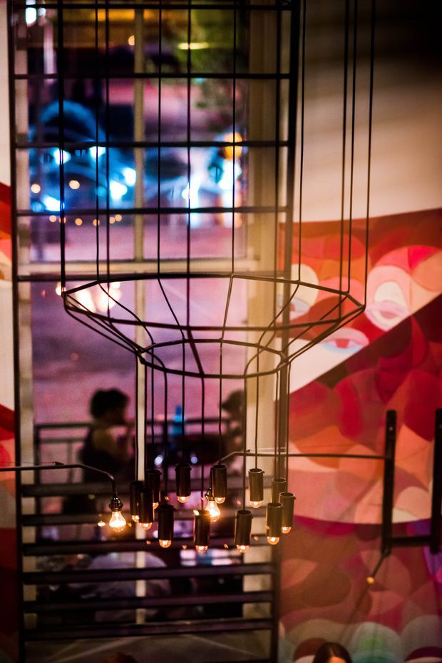 A dimly lit contemporary interior featuring a large sculptural black metal pendant chandelier with numerous exposed warm-glowing bulbs. Floor-to-ceiling gridded glazing or a metal-framed screen reveals blurred activity beyond, while a vivid abstract red, pink, orange and white wall mural adds a dramatic backdrop. The space appears hospitality or commercial in character, with reflective surfaces and layered ambient lighting.