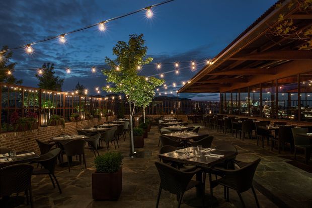 A warmly lit outdoor restaurant terrace at dusk, with multiple dining tables and dark woven chairs arranged across a paved courtyard. String lights create a festive canopy overhead, while a timber-and-glass restaurant pavilion runs along the right side. Brick planter walls, timber screening, potted plants, and a central tree define the landscaped dining area, with distant hills visible beyond.