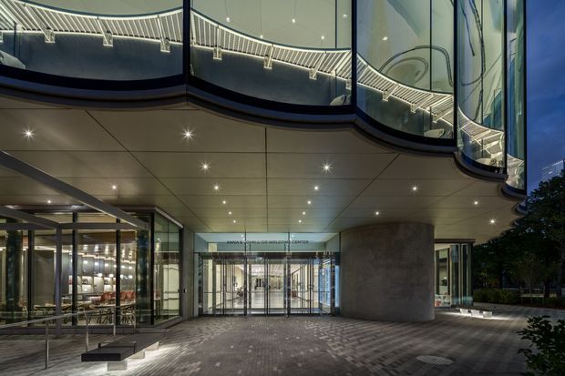 Night-time exterior view of a sophisticated commercial building entrance beneath a broad cantilevered canopy. The facade features extensive curved and angled glass, polished metal framing, recessed downlights, a large cylindrical concrete or stone-clad column, glazed entry doors, and patterned dark paving. A brightly lit interior is visible through the glazing.