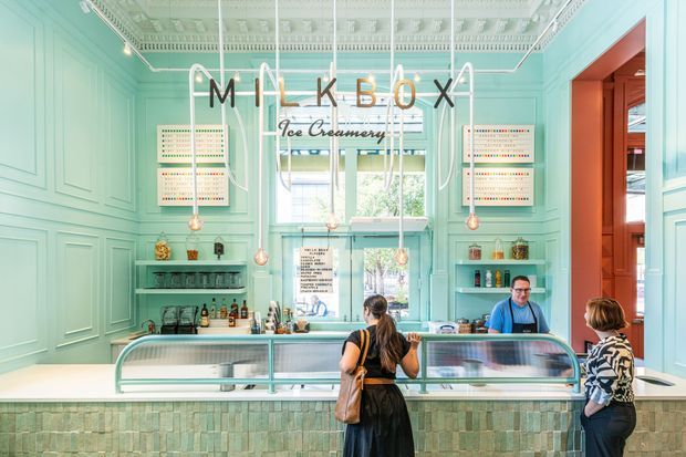 Bright, mint-green ice cream shop interior with ornate pressed ceiling detailing, decorative wall panelling, pendant lighting, open display counters, and a tiled service counter. Customers and staff occupy the foreground, with large front windows and a warm-toned doorway visible at the right.