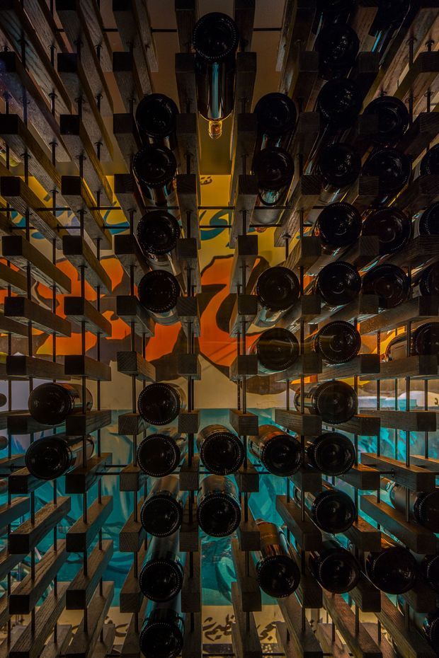 A dramatic interior wine cellar or wine display installation viewed through a dense grid of horizontally stored dark wine bottles. Timber or dark metal shelving creates a strong geometric perspective, with warm amber and orange lighting above and vivid turquoise-blue illuminated areas behind and below. The composition feels immersive, sculptural, and highly atmospheric, with repeated cylindrical bottle ends dominating the frame.