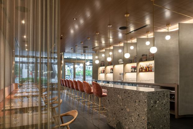 A refined contemporary commercial bar and dining space with a long terrazzo-faced counter, rust-coloured upholstered bar stools, dark timber ceiling, concrete-toned walls, illuminated bottle shelving, and a row of spherical pendant lights. A sheer chain curtain partially screens a dining area with timber tables and chairs on the left, while glazed openings provide views toward the exterior.