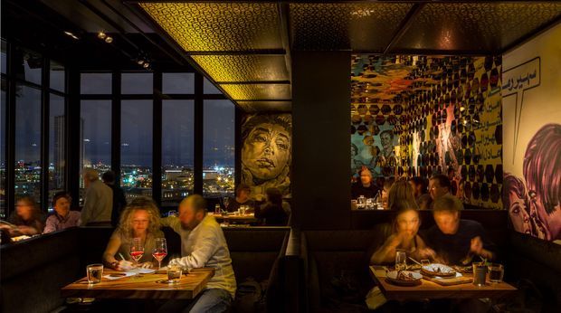 A dimly lit rooftop-style restaurant or bar interior with panoramic floor-to-ceiling windows overlooking a city at night. Dark upholstered booth seating and small dining tables fill the space, while patterned metallic ceiling panels, warm amber lighting, and colourful mural artwork create a dramatic, eclectic atmosphere.