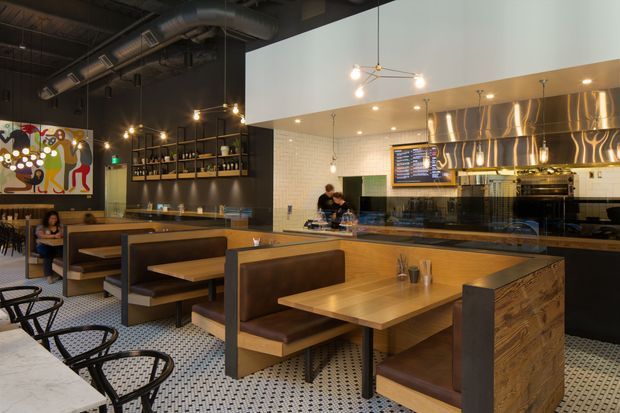 A lively modern restaurant interior with booth seating, communal tables, a service counter and open kitchen. The space combines warm timber and brown leather furnishings with a black ceiling, exposed mechanical services, pendant lighting, graphic artwork, patterned floor tiles, and a reflective metallic kitchen backsplash.
