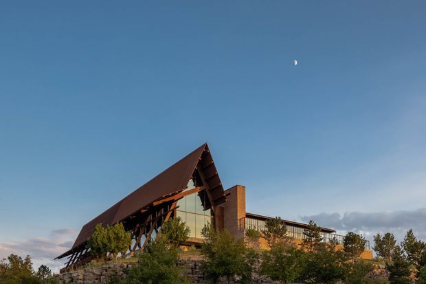Exterior architectural photograph of a large, low-rise building set on a landscaped, rocky site. The composition is dominated by a steeply pitched, asymmetrical gable roof with deep exposed rafters and dark brown metal cladding. A tall glazed gable end, stone retaining walls, warm brickwork, and timber or dark structural framing are visible. The building is surrounded by trees and shrubs under a clear blue sky, with a small crescent moon overhead.