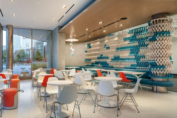 A bright contemporary commercial café or communal lounge with terrazzo flooring, extensive floor-to-ceiling glazing, pale stone walls, and a warm timber slatted ceiling. Numerous white round tables and molded chairs are arranged throughout the space, with vivid red and turquoise seating accents. A striking sculptural turquoise-and-white patterned feature wall and matching column define the back of the room.