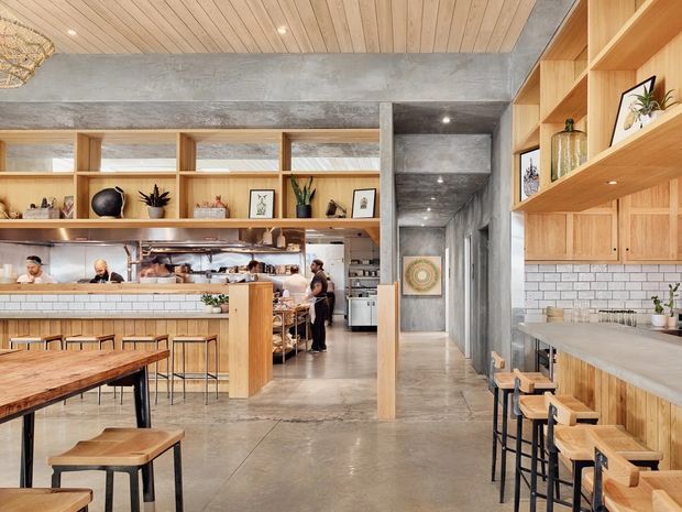 Contemporary café or restaurant interior with an open kitchen and service counter, communal timber tables, and a long built-in bar seating area. The space combines polished concrete floors and structural beams with warm timber cabinetry, open shelving, a pale tiled backsplash, and a slatted timber ceiling. Industrial-style black metal stool frames contrast with the warm wood fit-out, while plants and decorative objects add a refined hospitality atmosphere.