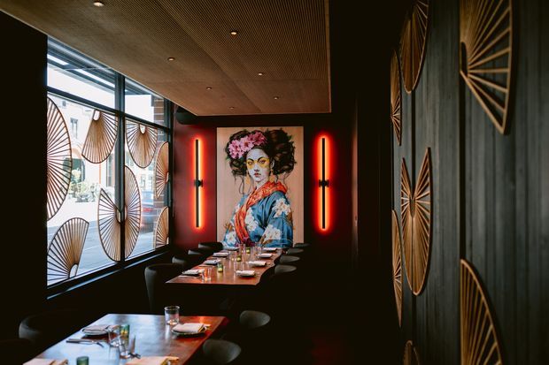 A moody, sophisticated restaurant dining interior with dark walls, booth and table seating, a timber slatted ceiling, dramatic red vertical lighting, decorative geometric wall panels, and a large colourful portrait artwork. Floor-to-ceiling glazing on the left features patterned screening and brings in daylight.