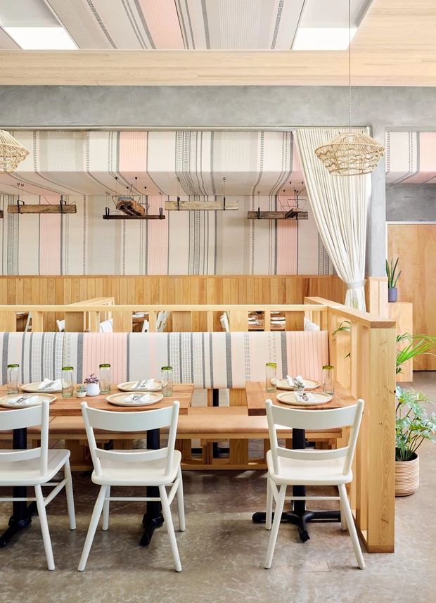 Bright contemporary restaurant dining interior with a long upholstered banquette, multiple light wood dining tables, white chairs, and patterned vertical upholstery extending onto the ceiling. The space features warm timber wall panelling and partitions, a raw concrete structural beam, hanging woven pendant lights, exposed suspended shelving, potted greenery, and a polished concrete floor.