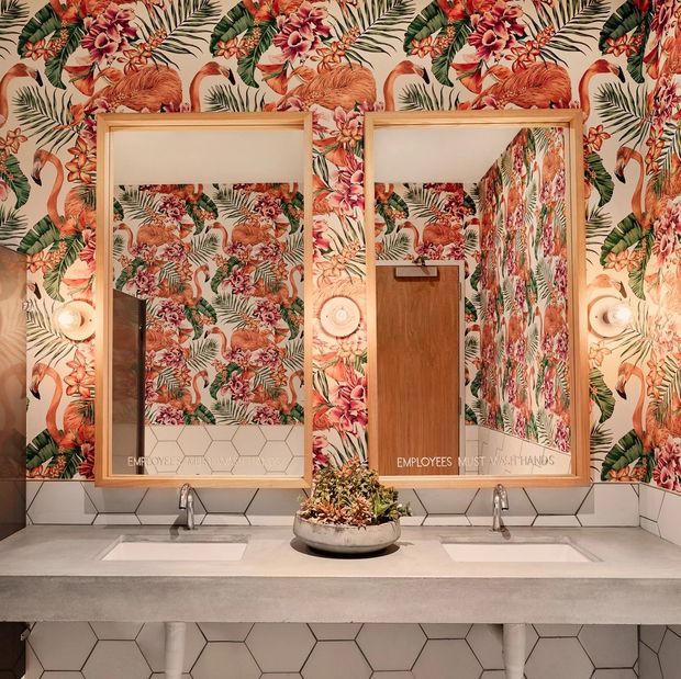 A stylish commercial bathroom vanity with two wall-mounted rectangular mirrors above a long concrete or stone counter with twin white basins and exposed chrome faucets. The walls and mirror interiors feature bold tropical floral wallpaper with flamingos, foliage, and pink-red flowers. White hexagonal tiles form the backsplash and lower wall, while warm timber mirror frames and a central planter add contrast. The mirrors include faint employee handwashing signage, suggesting a public or hospitality restroom.