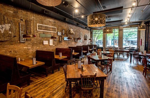 A warmly lit restaurant dining interior with exposed aged brick walls, dark timber ceiling and ductwork, polished timber flooring, numerous timber tables and chairs, and built-in booth seating. Decorative pendant lights, framed wall objects, and large front windows create an industrial yet inviting atmosphere.