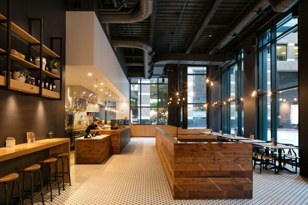 A stylish industrial café or restaurant interior with exposed ductwork and services, tall black-framed glazing, warm timber counters and bar seating, dark walls, and decorative shelving. The space features a central timber service island, patterned tile flooring, pendant and string lighting, and views to the urban street beyond.