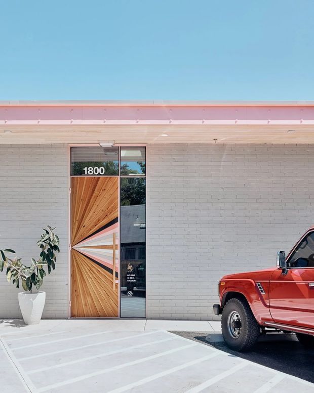 Exterior view of a low-rise commercial or residential building with a pale grey painted brick facade, a distinctive timber pivot entry door with a geometric fan pattern, and a narrow glazed sidelight marked “1800.” A red vintage utility vehicle is parked at the right edge, while a potted plant and striped accessible-style paving occupy the foreground. The building has a low-profile roof with a pale pink fascia and a clean, colourful contemporary facade.