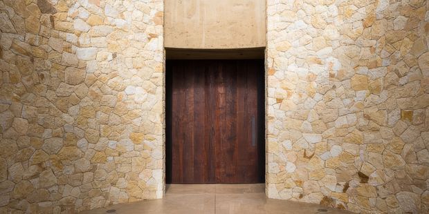 Close-up architectural view of a tall dark timber door recessed within a thick wall finished in irregular warm-toned natural stone. The stonework wraps around the doorway, creating a heavy, monolithic entrance with a simple tiled or stone floor at the base.