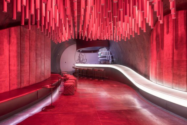 Dramatic futuristic interior lounge or bar with a predominantly red and magenta colour scheme. The space features curved walls and ceiling, a sculptural illuminated white counter or banquette sweeping through the room, small bar tables and stools, and a dense installation of suspended red cylindrical elements across the ceiling. The highly polished floor and integrated lighting create a bold, immersive, nightclub-like atmosphere.