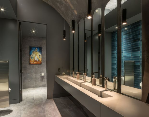 A dramatic contemporary bathroom vanity with a long pale stone counter and multiple integrated basins, brushed metal taps, and tall black-framed mirrors. The dark grey walls and concrete-look tiled floor create a moody, industrial atmosphere, while a glossy deep teal-blue tiled wall adds contrast. Black cylindrical pendant lights hang above the vanity, and an open doorway leads to a tiled shower or wet area.