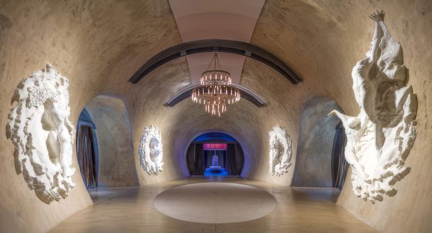 A dramatic, cave-like interior with rough sculpted beige walls, large white sculptural reliefs on both sides, curved dark ceiling beams, and a central contemporary chandelier. The symmetrical room leads toward a dark stage or alcove illuminated in blue and purple, with a circular pale floor feature in the foreground. The atmosphere is theatrical, immersive, and luxurious.