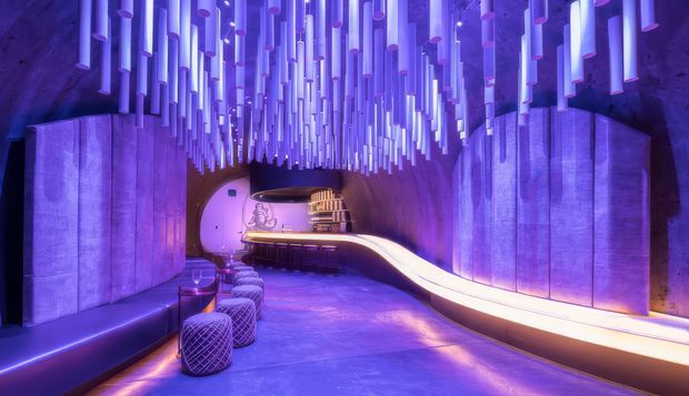 A futuristic, immersive commercial interior with a sweeping illuminated bar or reception counter, curved walls, built-in bench seating, and a dense suspended installation of glowing vertical tubes across the ceiling. The space is lit predominantly in violet, purple, and blue, creating a dramatic atmospheric lounge or hospitality setting.