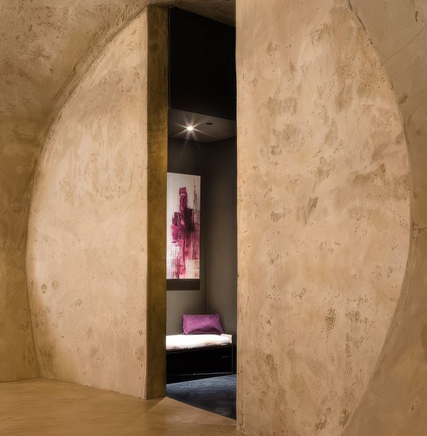 A narrow recessed doorway opens through sculptural, heavily textured beige plaster walls into a dark, intimate bedroom-like space. The alcove contains a low bed or daybed with white bedding and a vivid magenta cushion, dark flooring, a dramatic abstract artwork, and a small ceiling spotlight. The room has a warm, moody, tactile, minimalist character with monolithic curved wall forms.