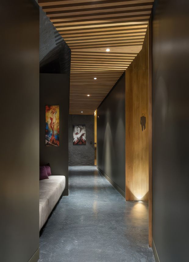 A long, narrow, dimly lit interior hallway with dark painted walls, a polished concrete floor, and a striking slatted timber ceiling with recessed downlights. Timber wall panelling and integrated uplighting frame the corridor, while a partial light-coloured sofa and colourful artwork are visible along the left side. The atmosphere is dramatic, warm, and contemporary.