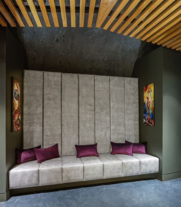 A dramatic, compact interior seating nook with a long built-in upholstered banquette beneath a sculptural dark textured wall. The high back is divided into vertical padded panels, with deep burgundy cushions and colourful narrow artworks on either side. Exposed timber ceiling battens create a strong linear pattern overhead, while dark green-grey walls and a blue-grey floor give the space a moody, refined character.