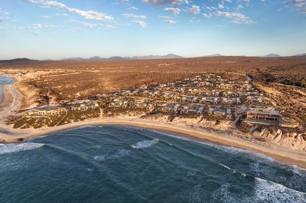 Aerial view of a coastal residential development spread across a rugged, sparsely vegetated shoreline, with clustered low-rise buildings set behind a sandy beach and ocean waves in the foreground. Arid terrain and distant mountains form the backdrop under a partly cloudy sky.