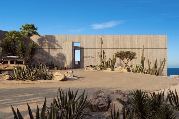 A minimalist single-storey coastal building with a long, monolithic tan concrete or rendered facade, narrow vertical entry opening, and sparse desert landscaping. Agave, yucca, cacti, boulders, and a sculptural tree surround a sandy forecourt, with ocean visible beyond the right side. The image has a clean architectural photography style under bright blue skies.