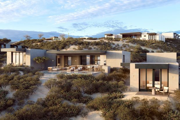 Wide exterior view of a low-profile contemporary residential development set among sparse coastal or desert-like native scrub. Multiple flat-roofed buildings use pale rendered walls, dark trim, expansive glazing and sheltered outdoor terraces, with sandy planting and a distant ocean or hazy horizon visible behind.
