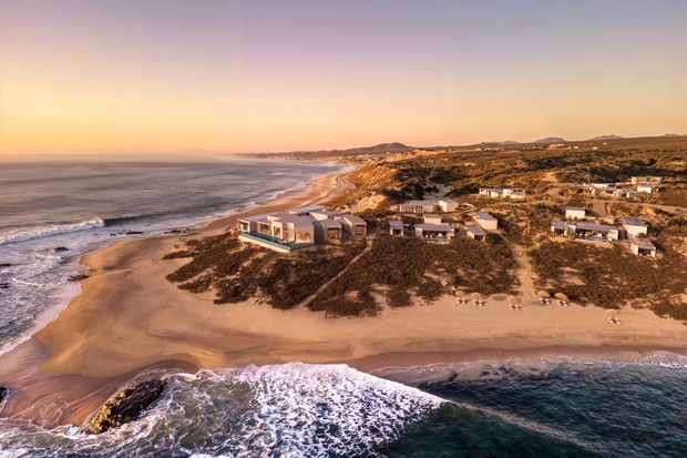 Aerial coastal view of a beachfront residential development set among sandy dunes, with several low-rise homes directly beside a broad beach and ocean shoreline. The scene is photographed in warm golden-hour light, with visible surf, sandy terrain and sparse coastal vegetation.