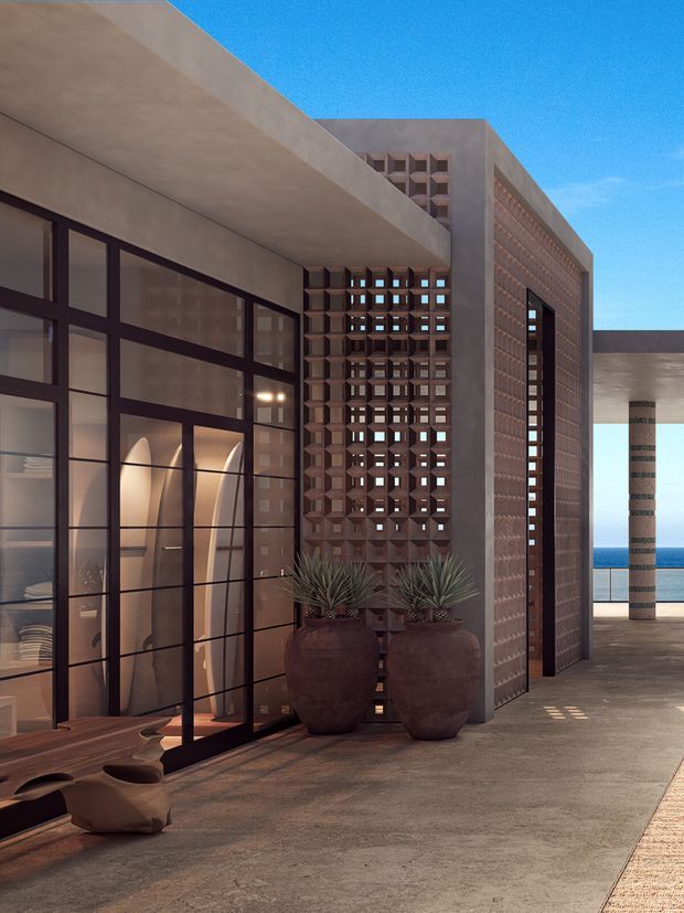 A contemporary exterior architectural view featuring a covered outdoor entry or terrace, large dark-framed glazing, exposed concrete walls and soffits, and a patterned terracotta breeze-block screen. Large ceramic planters and sculptural seating occupy the foreground, while an ocean horizon is visible beyond the building.