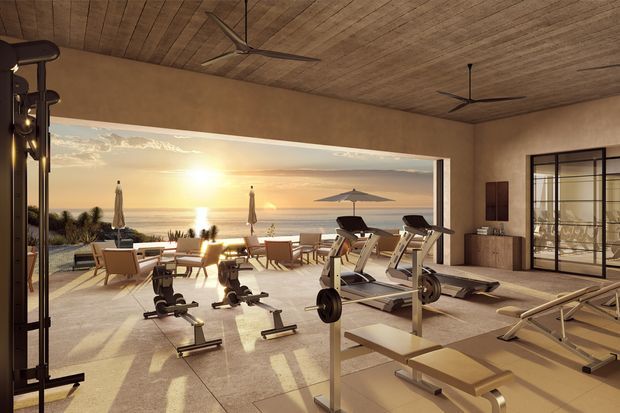A spacious coastal home gym with floor-to-ceiling open glazing overlooking a beach and ocean at sunset. The room contains multiple exercise machines, benches, free weights, and cardio equipment. Warm plaster walls, a wood-lined ceiling, ceiling fans, large tiled flooring, and dark-framed glazed doors create a refined resort-like setting.