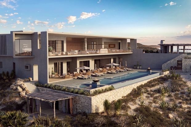A large contemporary multi-level residence set in a dry, rocky coastal or desert-like landscape. The building has crisp rendered and stone-clad walls, extensive open terraces, timber-look soffits, broad glazing, and a long rectangular swimming pool surrounded by sun loungers. A lower pergola and landscaped planting occupy the foreground, while a covered outdoor structure is visible to the right.