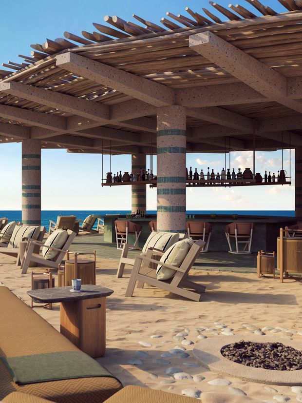 Open-air resort-style beachfront lounge beneath a broad timber pergola. The space includes sandy flooring with pebble detailing, timber-framed lounge chairs, low tables, a bar counter with hanging bottle display, striped structural columns, and unobstructed ocean views.