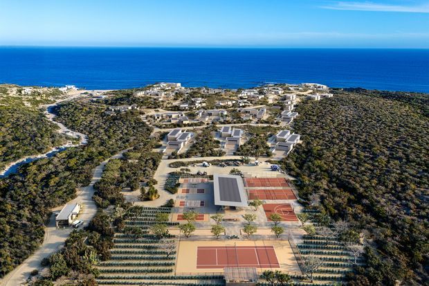 Aerial view of a coastal residential development set within dense natural vegetation, with multiple low-rise buildings, winding access roads, landscaped areas, and a prominent tennis and recreational court complex in the foreground. The deep blue ocean forms the background.