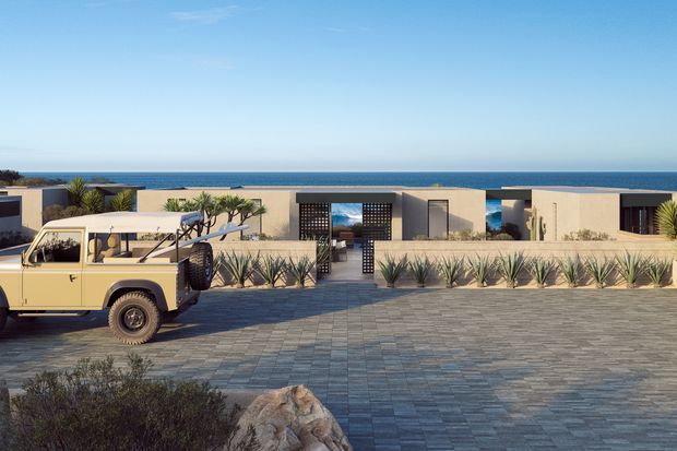 Wide exterior view of a low, contemporary coastal residence overlooking the ocean. The flat-roofed, beige-toned building is arranged around a paved forecourt with drought-tolerant landscaping and agave-like plants. A vintage utility vehicle is parked in the foreground, while the central entrance features a dark patterned screen and glazing. The image has a clean, modern resort-like character with strong horizontal lines and expansive ocean views.