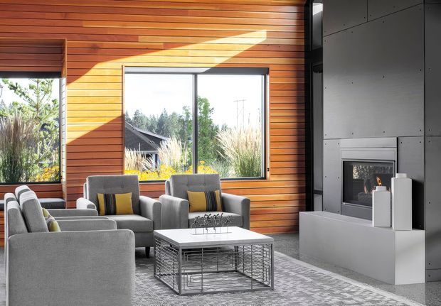 Bright contemporary living room with a dramatic timber-lined wall and large windows overlooking landscaped greenery. Several grey upholstered lounge chairs surround a rectangular glass-topped coffee table with a metal mesh base, while a dark grey paneled feature wall contains a modern built-in fireplace and low hearth. Sunlight creates a strong diagonal highlight across the timber wall.