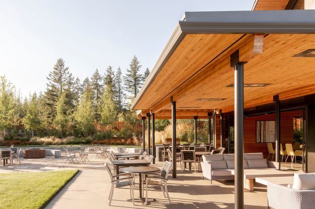Exterior view of a contemporary pavilion-style home with a broad flat roof and deep timber-lined soffit extending over a covered outdoor living and dining terrace. Slim dark structural posts support the canopy, while glazed and timber-clad walls open onto a paved patio furnished with outdoor sofas, dining tables and chairs. A landscaped garden with mature trees surrounds the terrace and includes a low circular fire pit or seating feature.