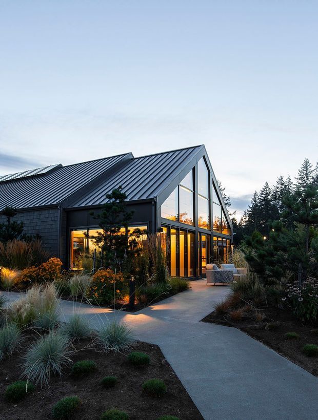 Exterior view of a contemporary gabled building at dusk, featuring a steep standing-seam metal roof, dark cladding, and extensive floor-to-ceiling glazing. Warm interior lighting glows through the glass, while a concrete pathway leads through a carefully designed garden with ornamental grasses, shrubs, and small trees toward an outdoor seating area.