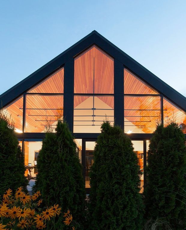 Front exterior view of a contemporary gable-roofed building at dusk. The facade combines dark structural framing with extensive floor-to-gable glazing and warm vertical timber lining visible beneath the roof. Dense evergreen screening and flowering garden plants occupy the foreground, while warm interior lighting creates a welcoming glow.