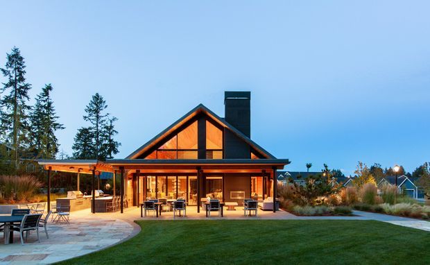 Exterior view of a warmly lit single-storey gable-roof home at dusk, featuring extensive timber-framed glazing, a deep covered outdoor entertaining area, stone-paved terrace, manicured lawn, landscaped planting, and a dark chimney or flue rising above the roof.