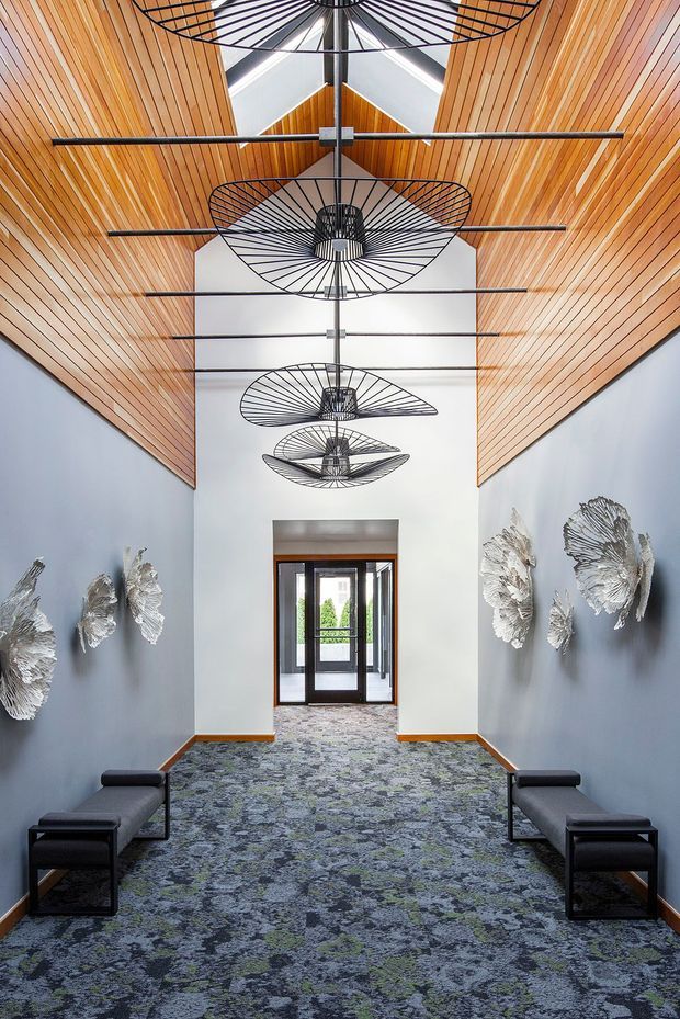 A long, narrow contemporary interior corridor or lobby with tall white walls, a dramatic pitched timber-lined ceiling, and repeated sculptural black wire pendant lights. Dark upholstered benches line both sides, while silver sculptural wall artworks decorate the blue-grey walls. Patterned carpet leads toward glazed timber-framed doors at the far end, creating a refined institutional or commercial entry space.