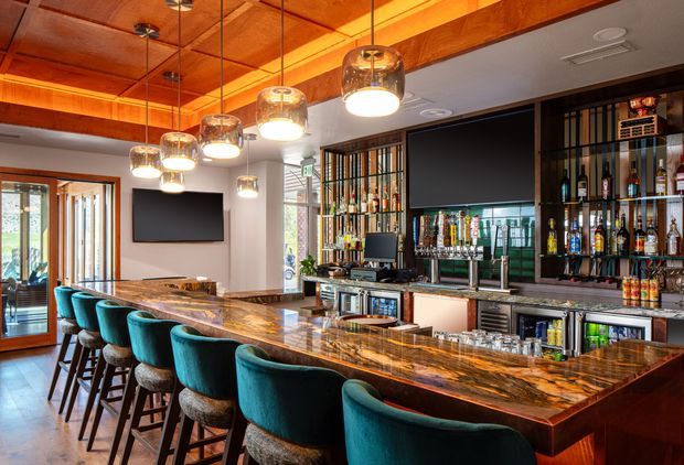 A sophisticated residential home bar with a long polished stone bar counter and seating for multiple people on teal upholstered stools. The room features extensive built-in bottle display and storage cabinetry, a central bar workstation, dark-framed glazing, warm timber ceiling panelling and beams, and a row of contemporary pendant lights. A television is mounted on the wall, with a bright glazed doorway at the side.